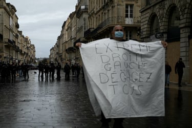 A masked protester holds a banner reading “far right, far left, get lost all of you” facing police