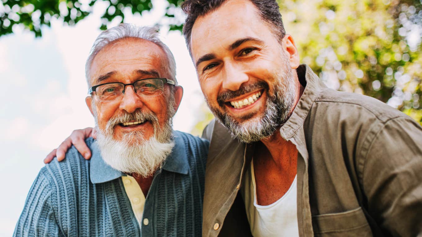 Older man with a white beard and glasses smiling beside an adult man with a beard, who has an arm around his shoulder, outdoors