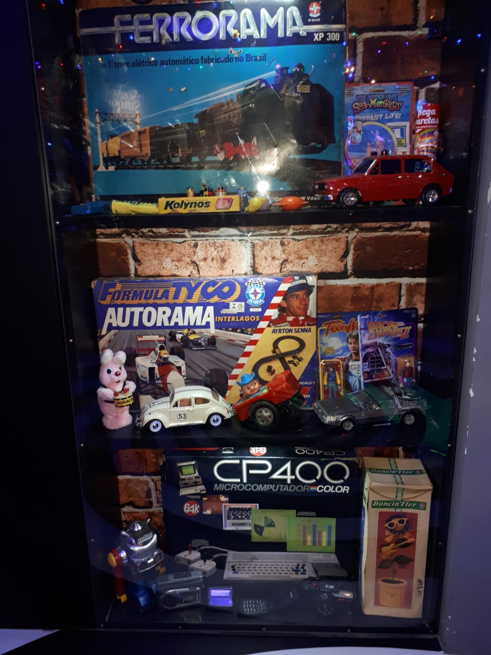 Uma estante cheia de objetos e brinquedos dos anos 80 e 90 está alocada em um canto.