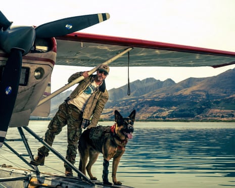 Brad Pitt stands on a floatplane's pontoon beside a German shepherd, lake and mountains behind