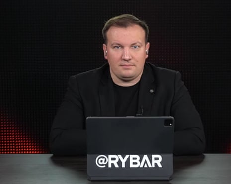 Mikhail Zvinchuk sits against a black background with an open laptop in front of him that has big white letters on the back of its screen spelling out Rybar. He wears earpieces as if he is taking part in a conference call, and is wearing a black jacket and top. He has short fair hair and is in his mid-30s.